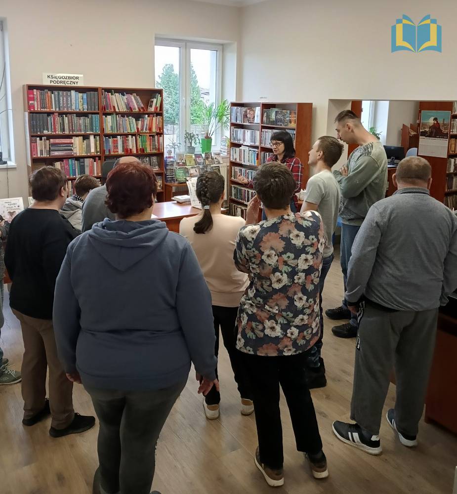 Zdjęcie: Bibliotekarka oprowadza dorosłych uczestników Warsztatów Terapii Zajęciowej po bibliotece.